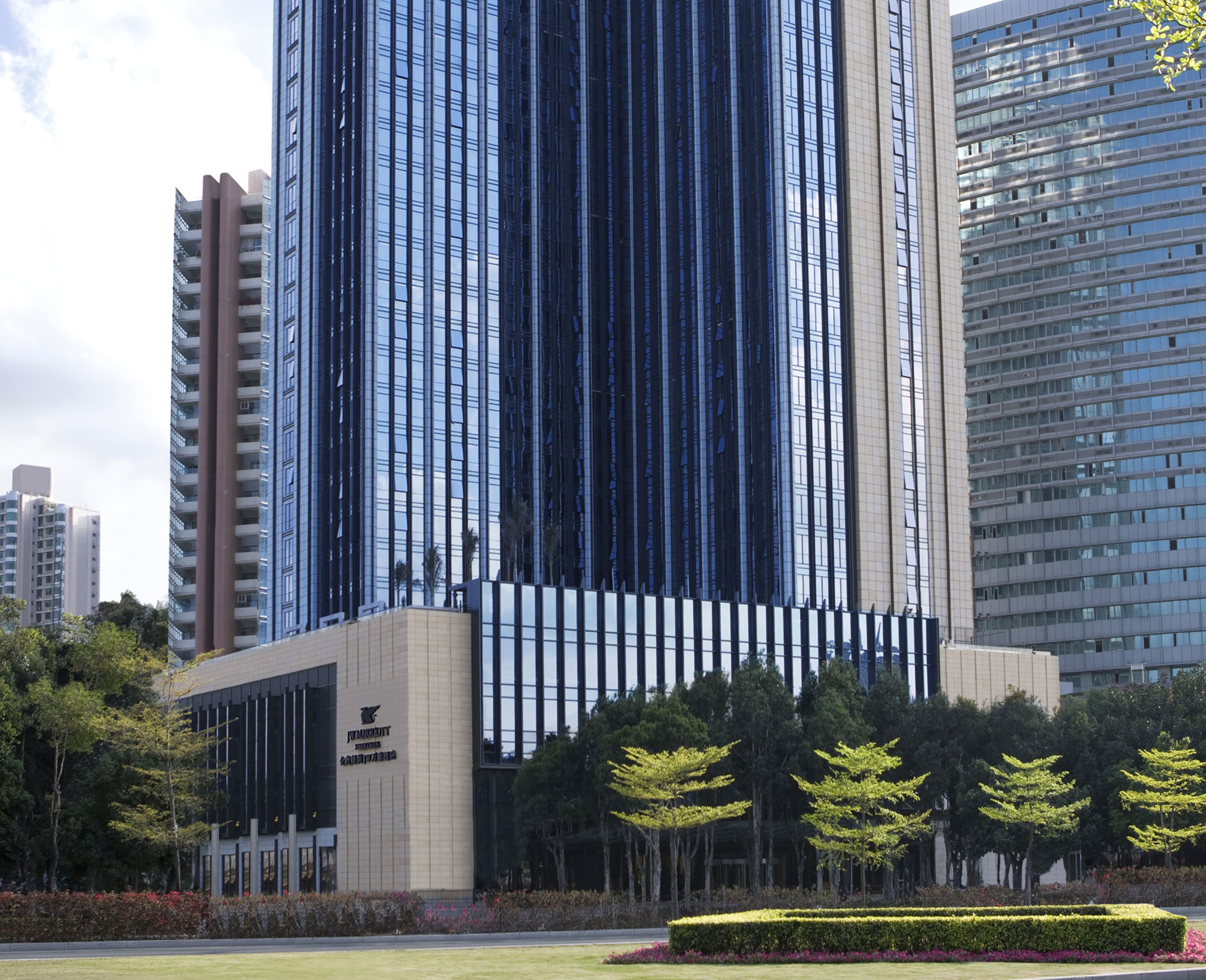 A modern high-rise building with reflective glass windows, surrounded by a well-maintained garden with neatly trimmed hedges and trees.