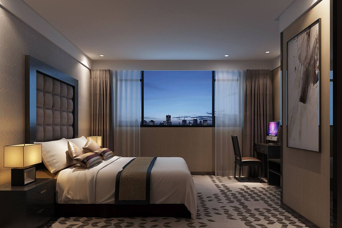 A modern hotel room with a large bed, a desk with a laptop, a city view, and a patterned carpet.