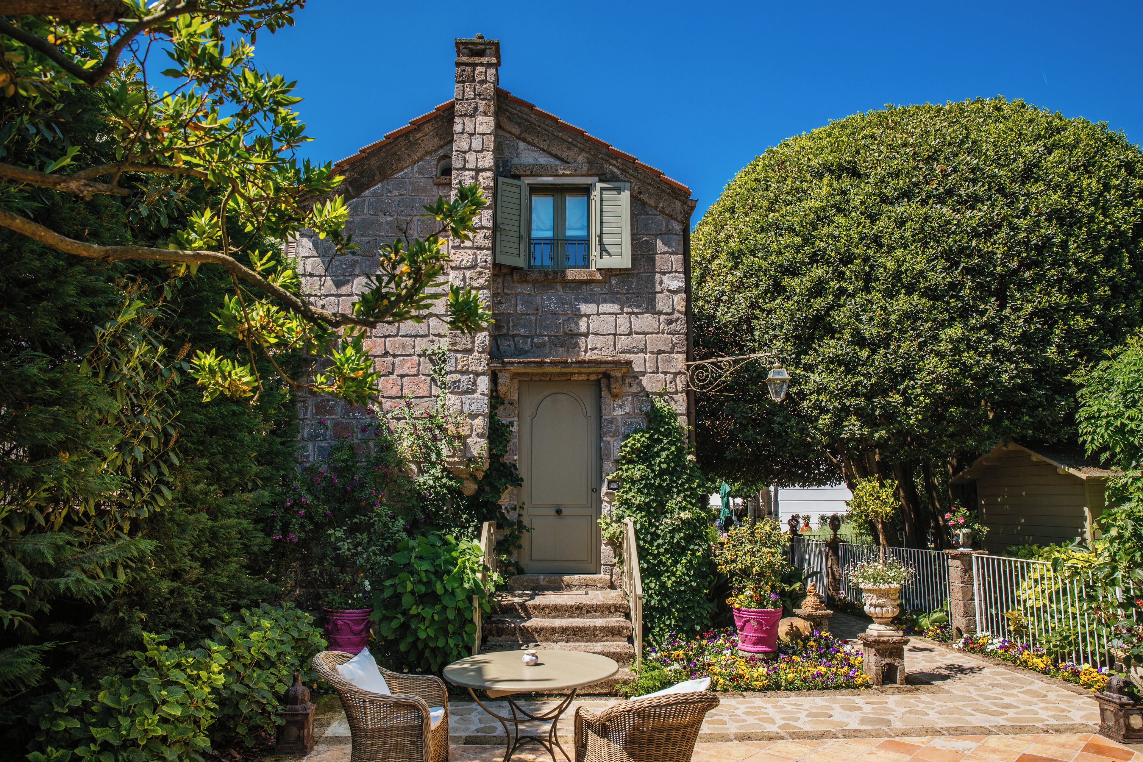 A stone house with a door and windows, surrounded by a garden with potted plants and a table with chairs.