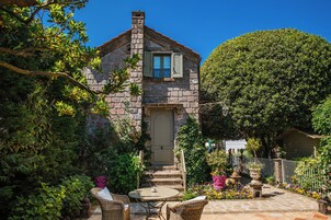 Una casa de piedra con puerta y ventanas, rodeada por un jardín con plantas en macetas y una mesa con sillas.