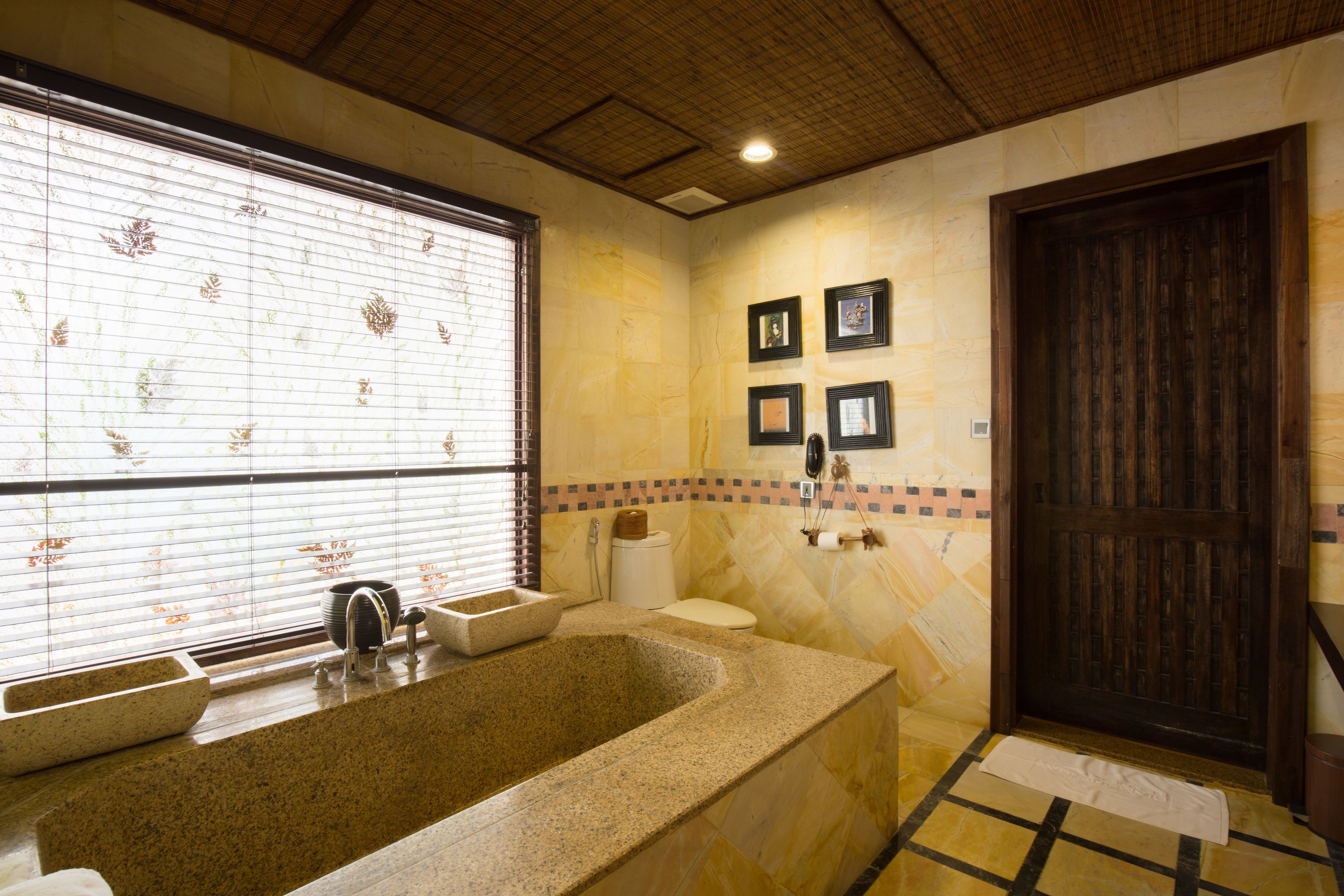 A bathroom with a large bathtub, a sink, and a toilet. There are framed pictures on the wall and a window with blinds.