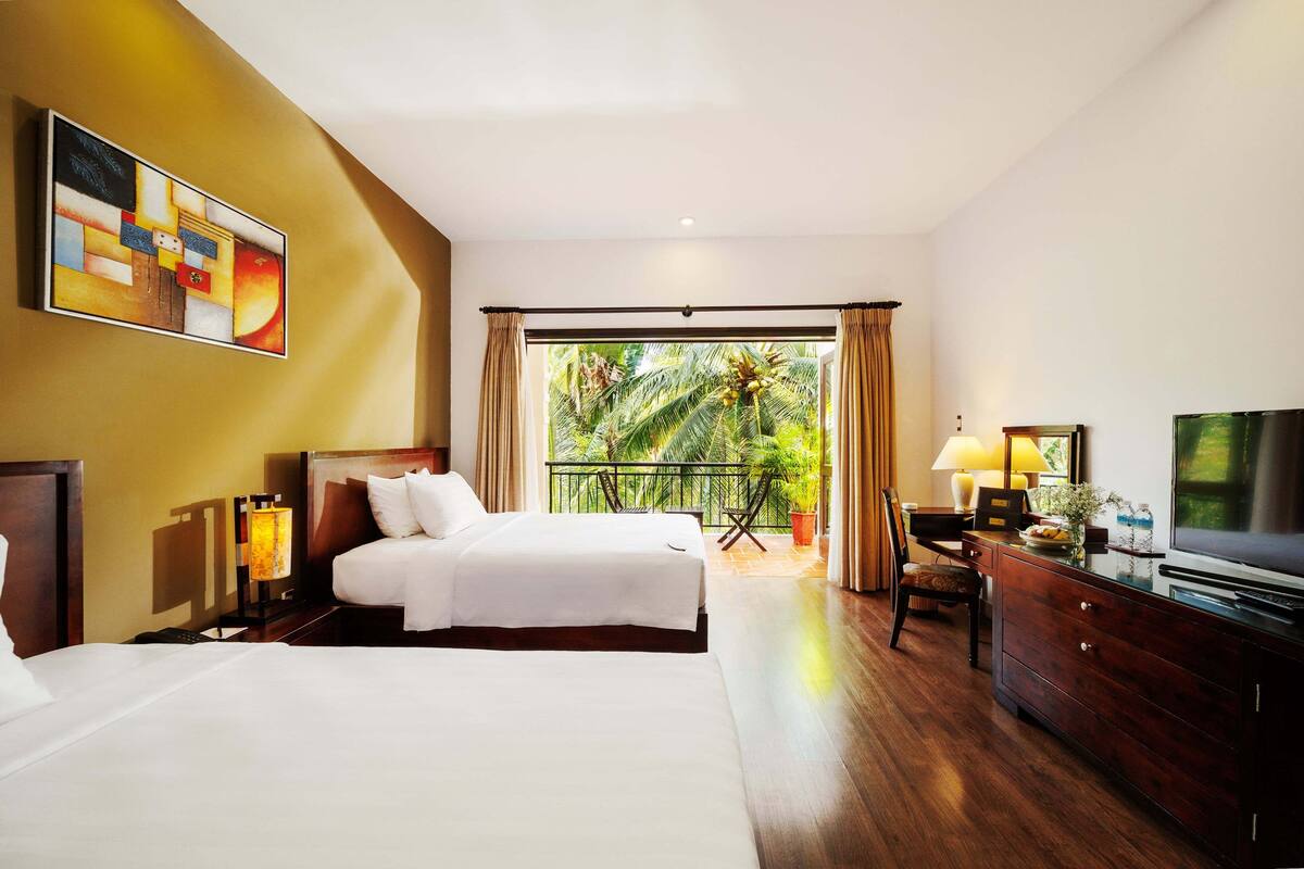 A hotel room with two beds, a TV, a desk, and a balcony with a view of palm trees.