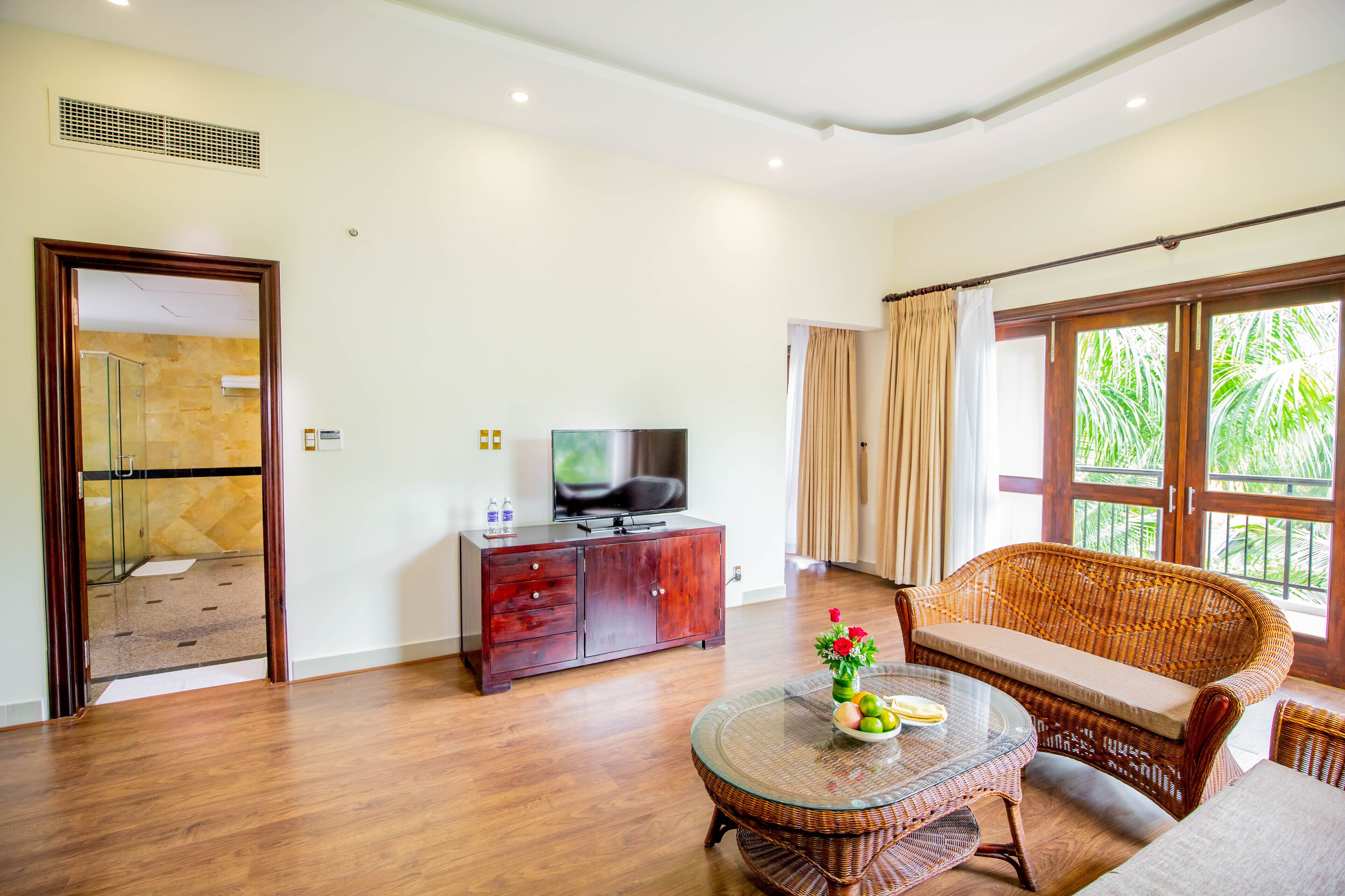 Un salón espacioso con suelo de madera, una mesa de centro de cristal con frutas, un sofá de mimbre, un televisor sobre un mueble de madera y un amplio ventanal con cortinas.