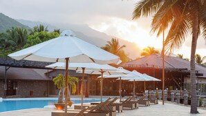 A poolside area with lounge chairs and umbrellas, a small palm tree, and a building with a tiled roof in the background. - Diamond Bay Resort and Spa (Nam Nha Trang)