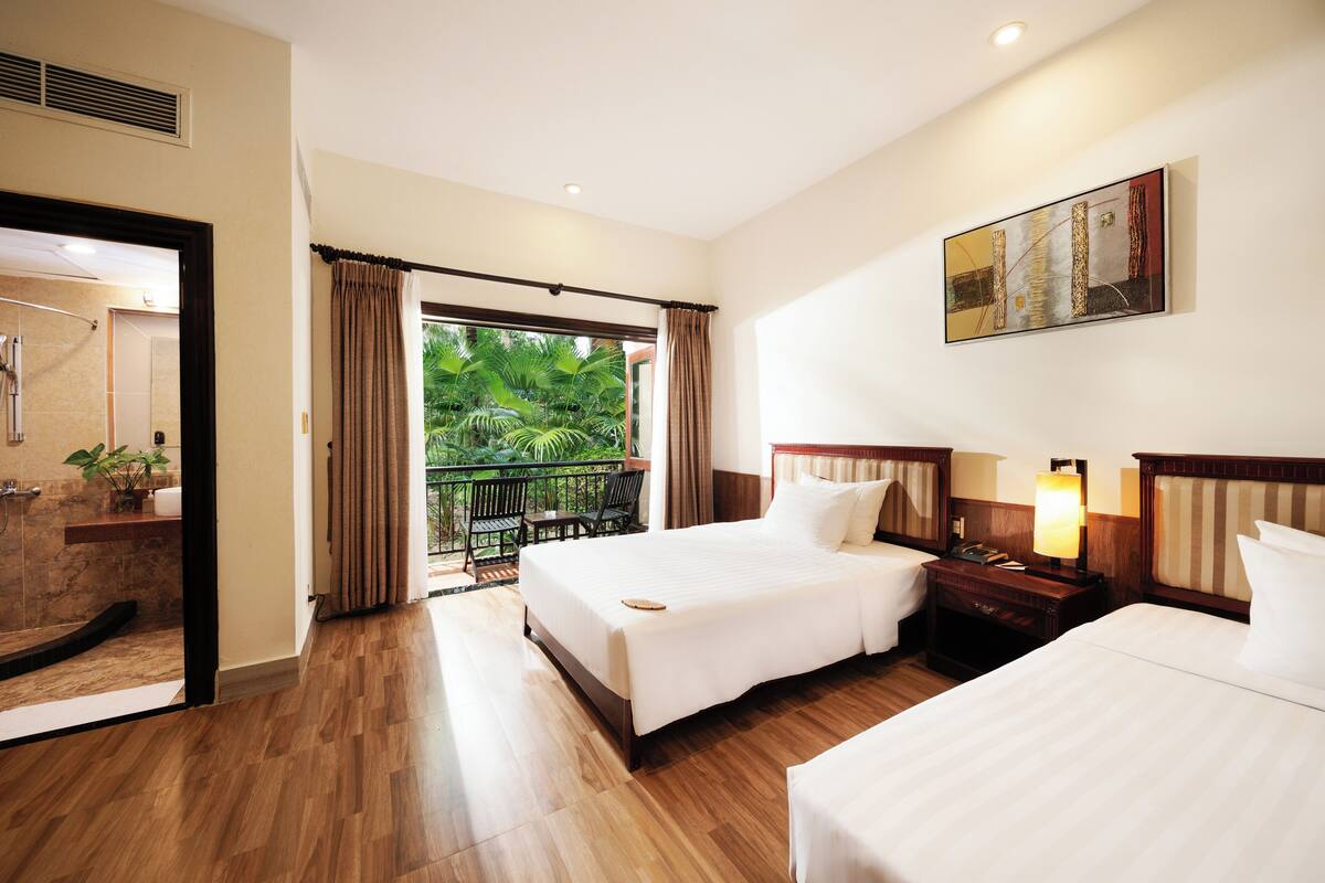 A hotel room with two beds, a balcony with a view of greenery, a bathroom with a shower, and a painting on the wall.