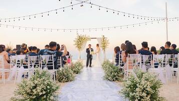 A beach wedding ceremony with guests seated on white chairs, a white aisle, and string lights overhead.