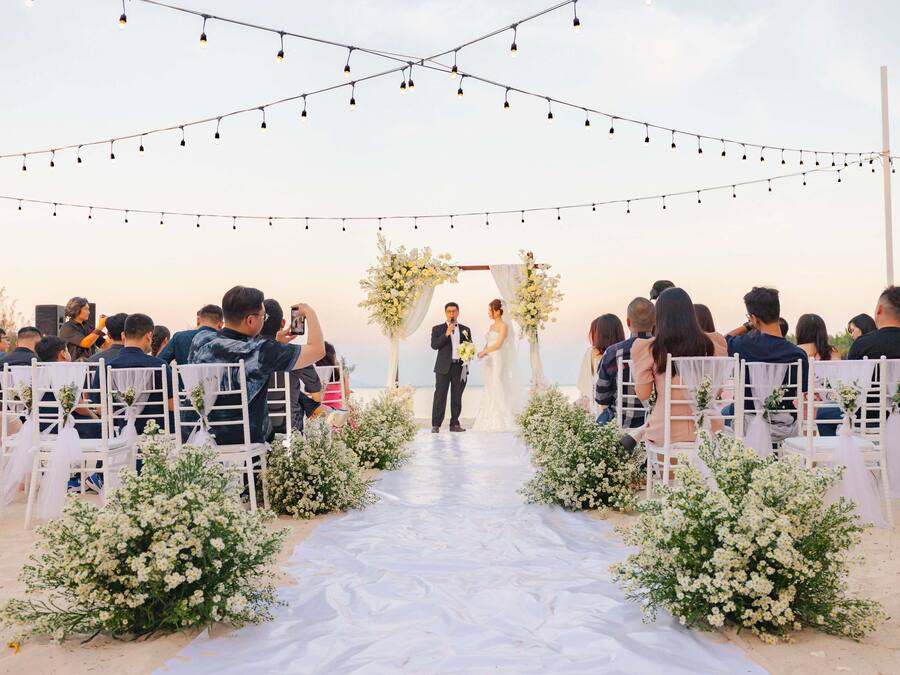 A beach wedding ceremony with guests seated on white chairs, a white aisle, and string lights overhead.