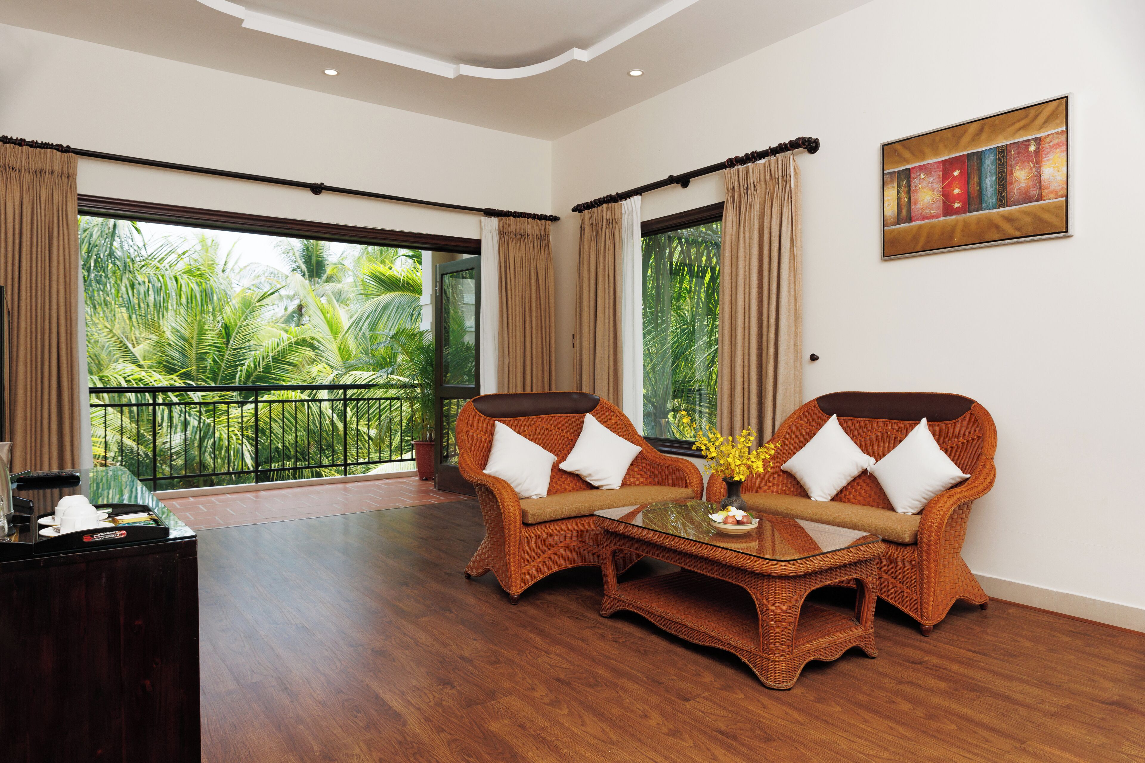 A room with a large window, a wicker sofa set, a small table with a vase, and a dark wooden desk.