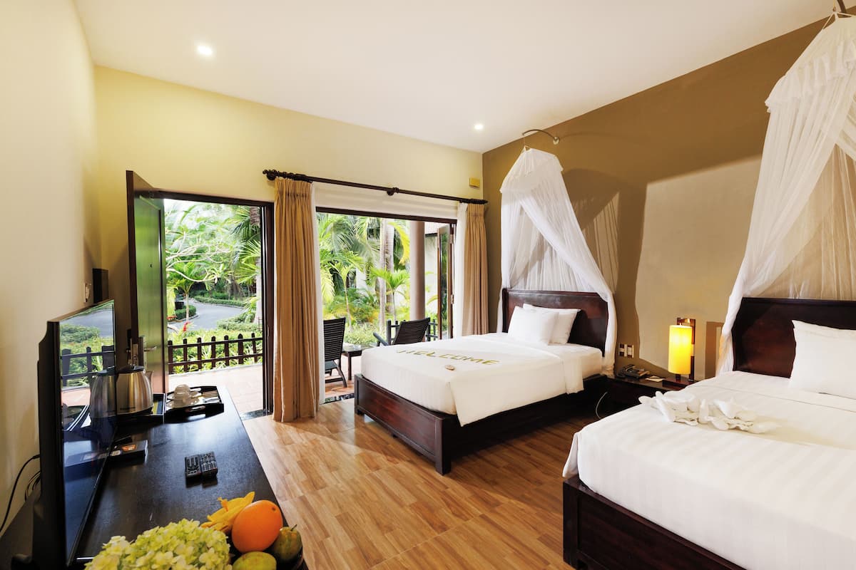 A hotel room with a bed, a desk with a computer, a TV, and a balcony with a view of greenery.