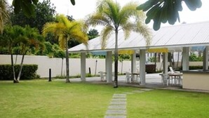 A covered outdoor seating area with tables and chairs, surrounded by a well-maintained lawn and palm trees. - Pacific Bay Hotel (Tamuning)