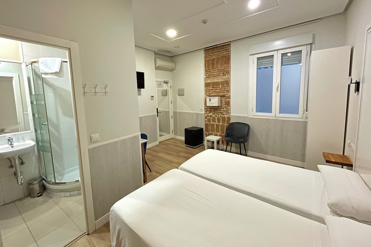 A hotel room with a bed, a small table, a chair, a bathroom with a sink and shower, and a brick accent wall.