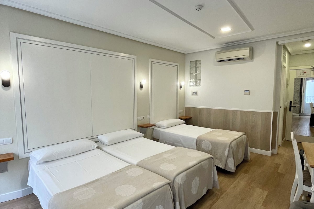 A hotel room with three beds, a wooden floor, an air conditioning unit, and a small dining area.