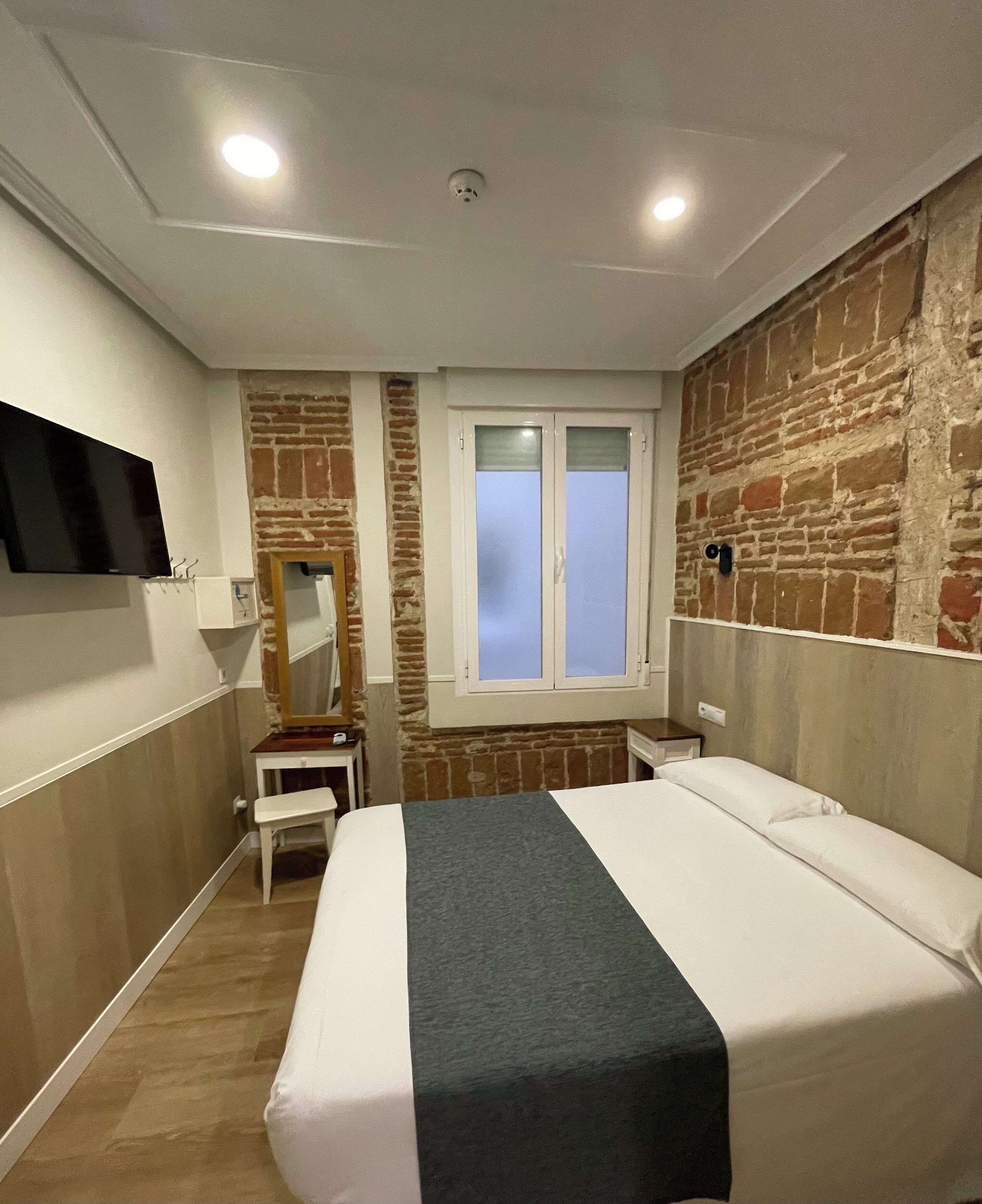 A hotel room with a brick wall, a window with frosted glass, a flat-screen TV mounted on the wall, and a bed with a grey bedspread.