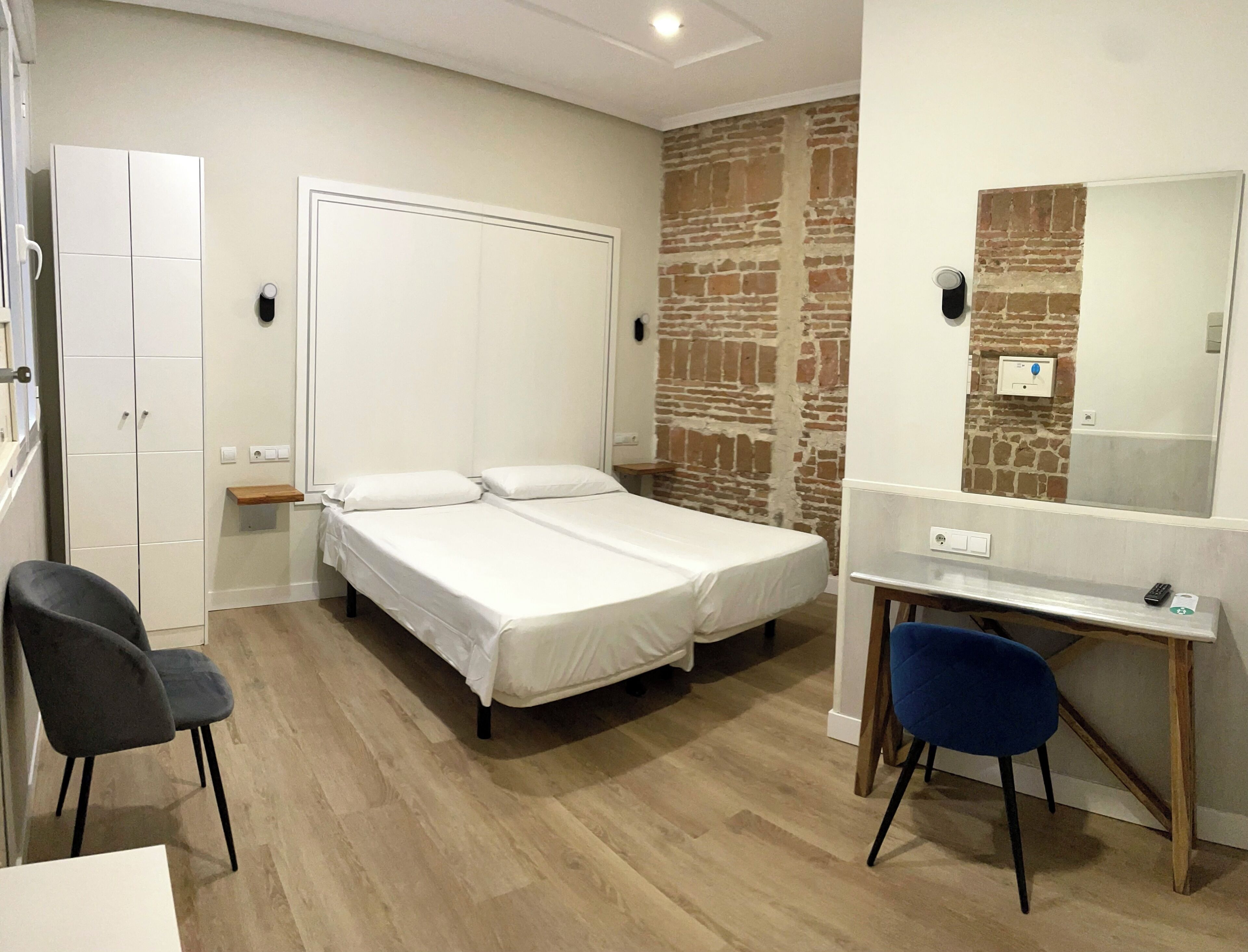 A room with two single beds, a desk with a chair, a wardrobe, and a brick wall.