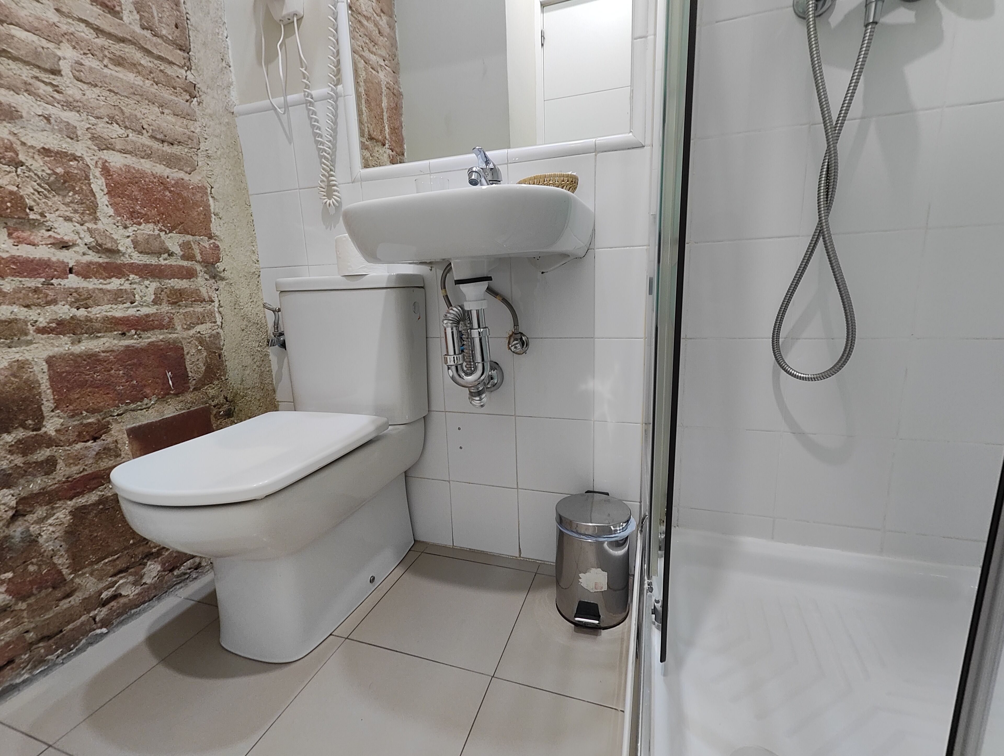Baño con inodoro blanco, lavamanos y ducha. Hay un cubo de basura y una pared de ladrillo.
