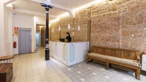 A reception area with a receptionist, a wooden bench, a tiled floor, and a brick wall. - Hostal Madrid (Madrid)