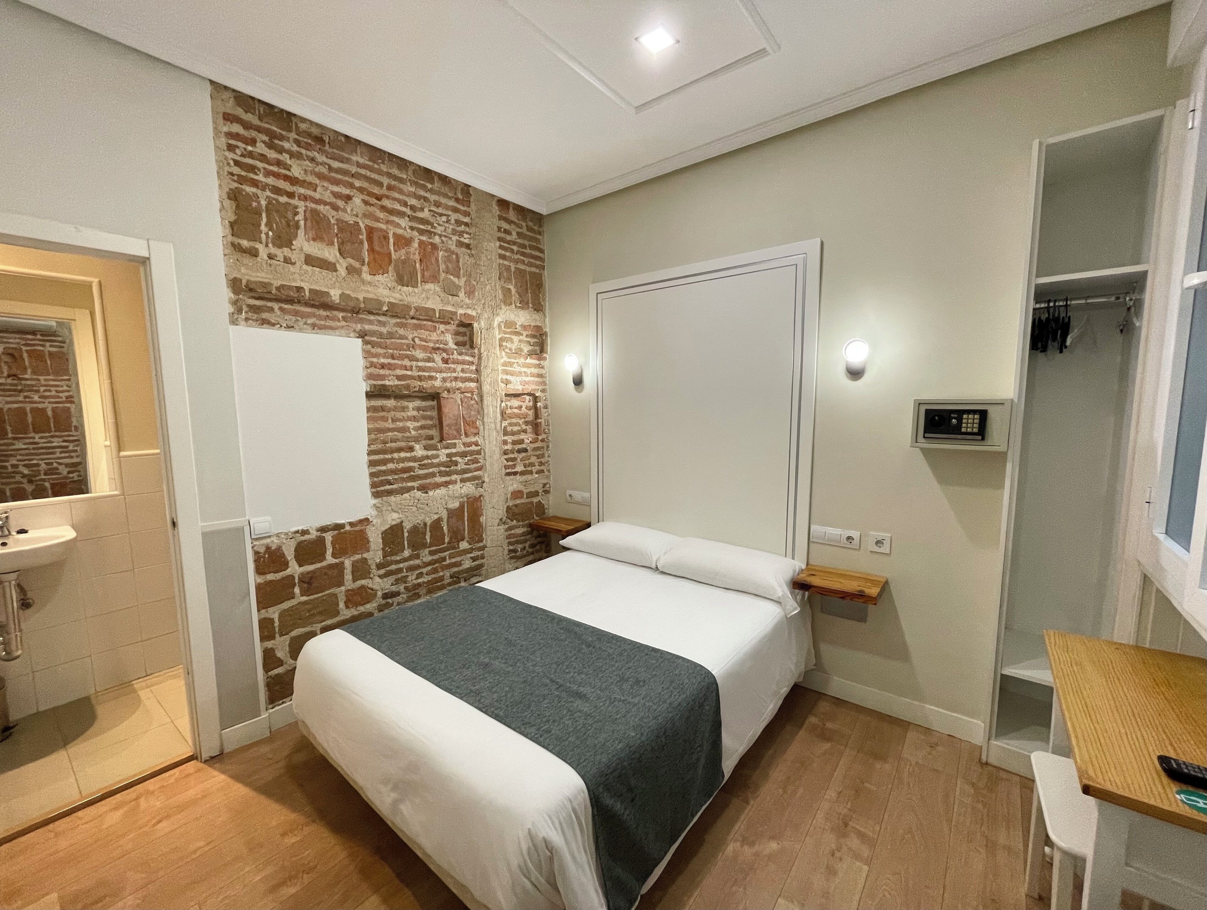 Un dormitorio con pared de ladrillo, una cama con ropa de cama blanca, una mesita de noche, un espejo, suelo de madera y un armario con ropa colgada.