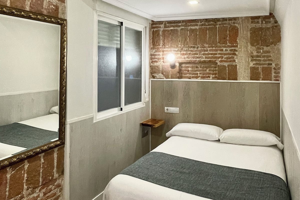 A bedroom with a brick wall, a large mirror, a bed with white and gray bedding, a small bedside table, and a window.