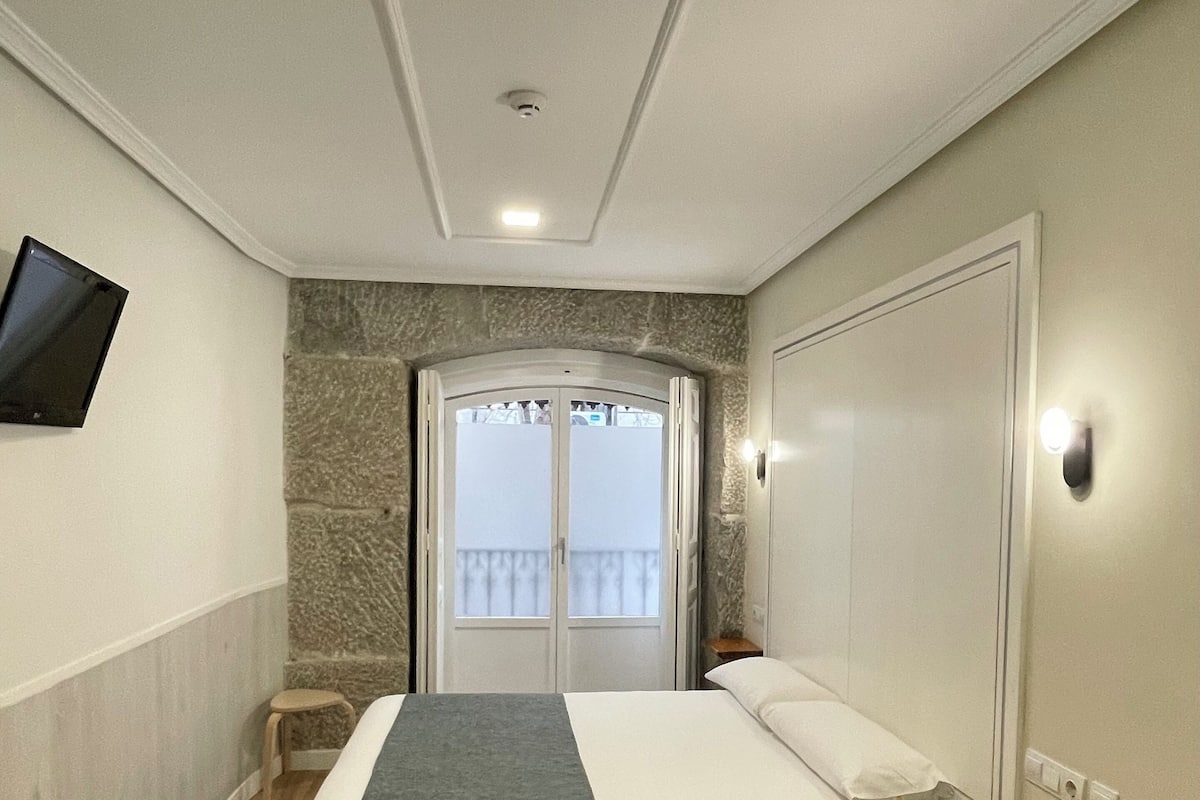 A bedroom with a bed, a bedside table, a TV mounted on the wall, and a door leading to a balcony.