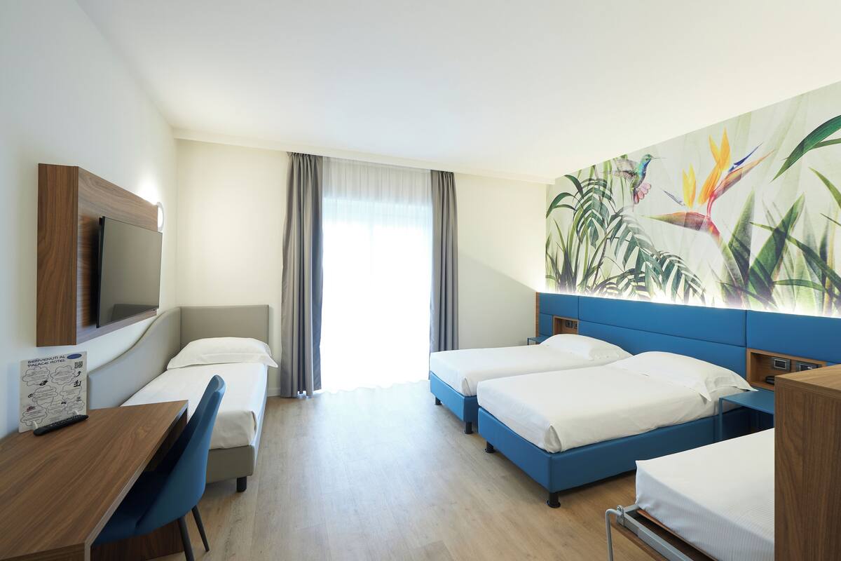 Hotellihuone, jossa on kaksi sänkyä, työpöytä, tuoli, televisio ja suuri ikkuna, jossa on verhot.