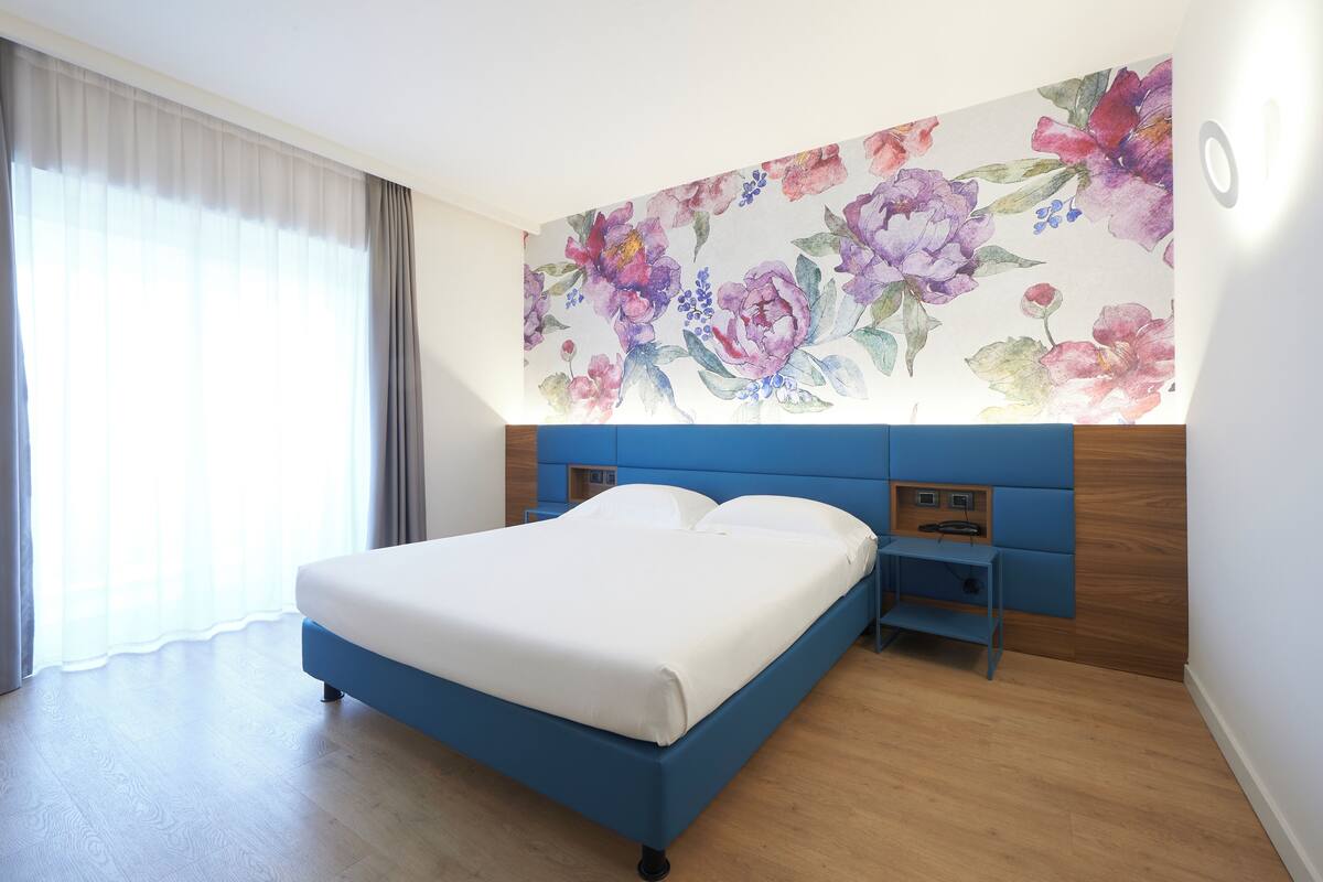 Hotellihuone, jossa on suuri sänky, puinen pääty, yöpöytä ja kukka-aiheinen seinämaalaus.