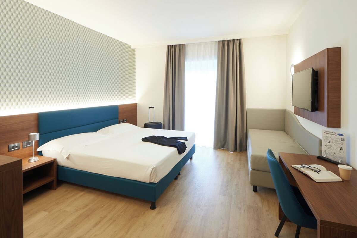 Hotellihuone, jossa on sänky, työpöytä, tuoli, televisio ja ikkuna, jossa on verhot.