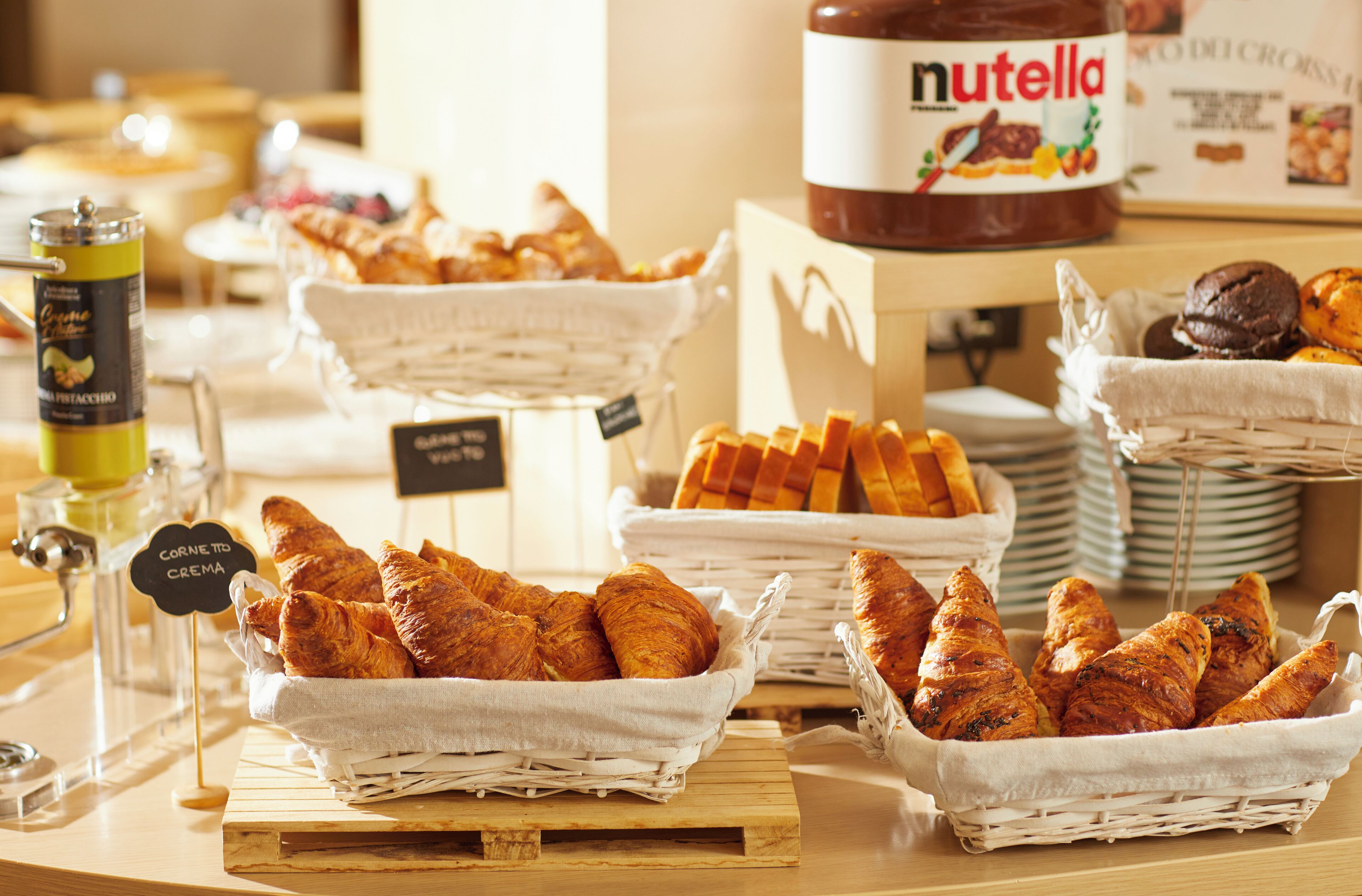 Un bufé de desayuno con cestas de cruasanes y otros pasteles, un tarro de Nutella y un dispensador de café.