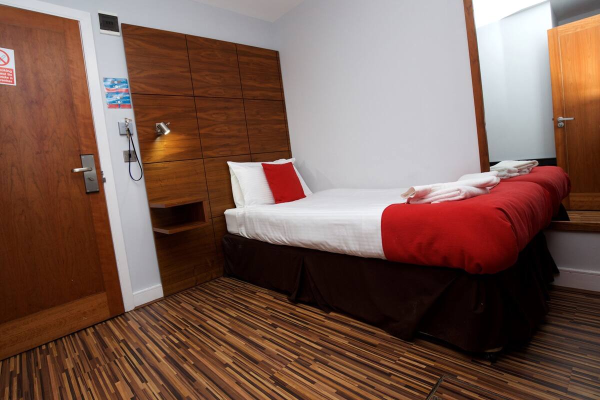 A hotel room with a wooden floor, a bed with white and red bedding, a wooden headboard, and a mirror on the wall.