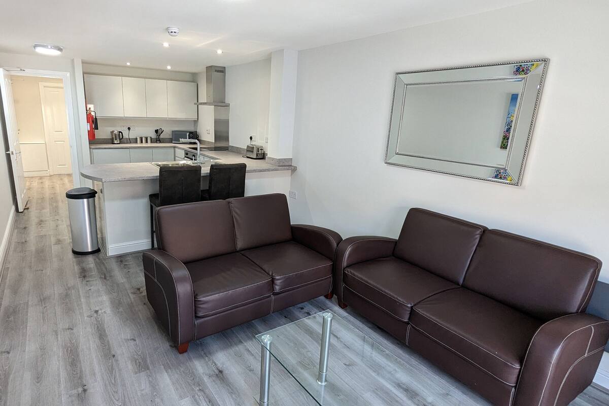 A modern living room with a brown leather sofa, a glass coffee table, and a kitchen in the background.