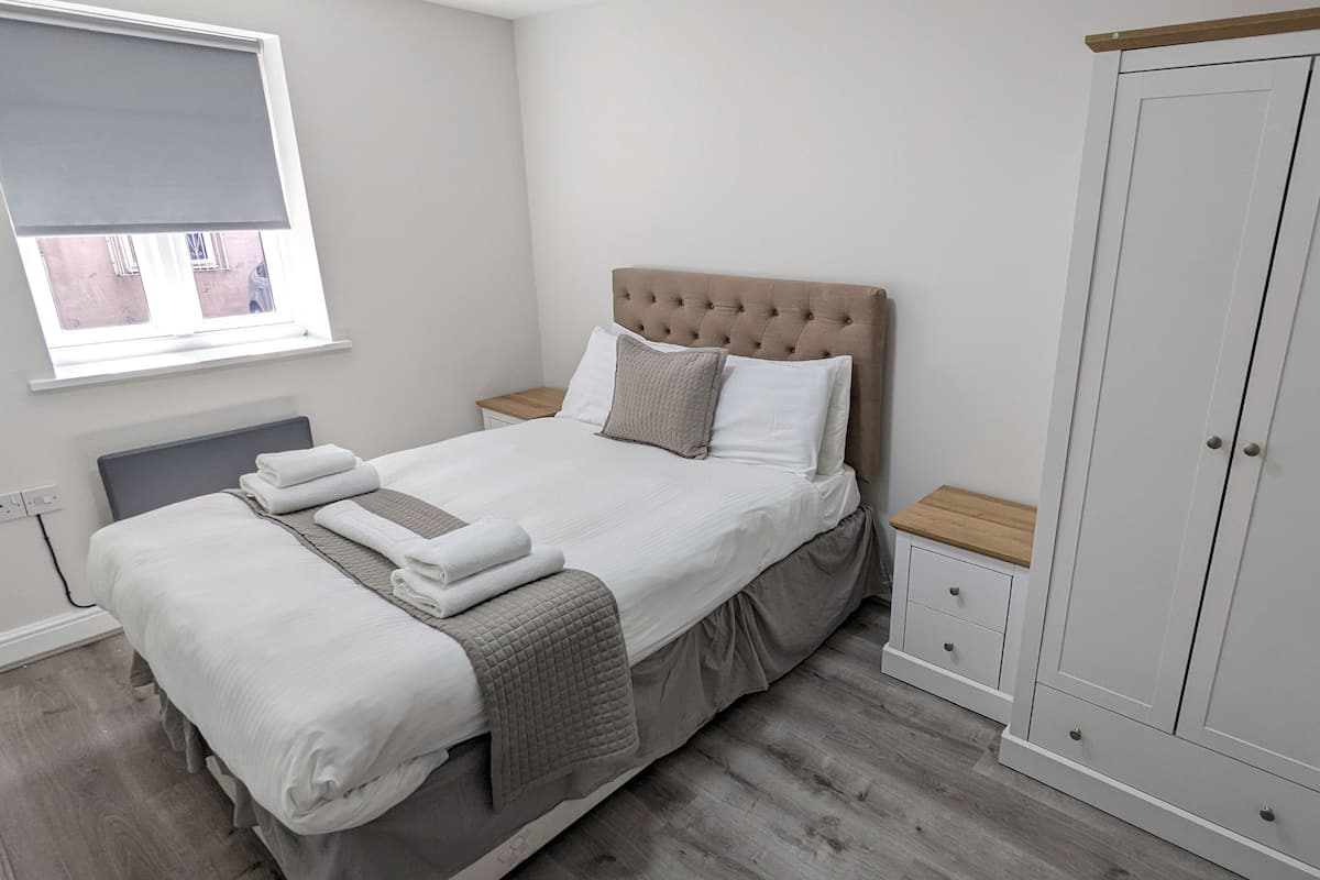 A neatly made bed with pillows and folded towels, a bedside table, a wardrobe, and a window with blinds.
