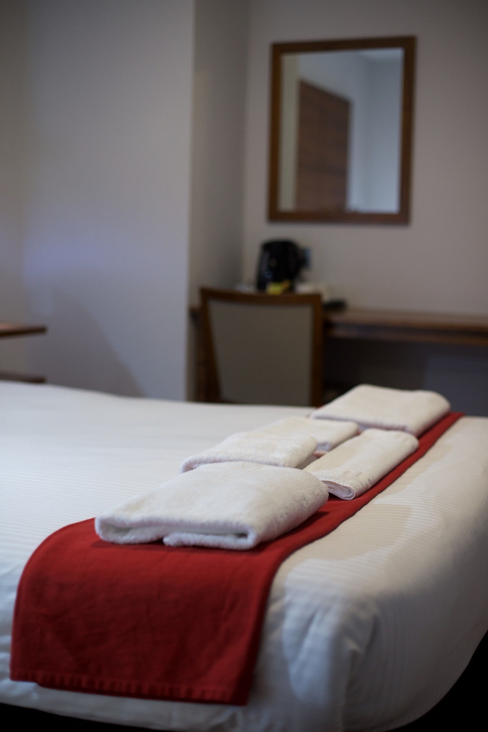 Un lit bien fait, avec du linge de lit blanc et une couverture rouge, un bureau en bois avec une chaise, et un miroir au mur.