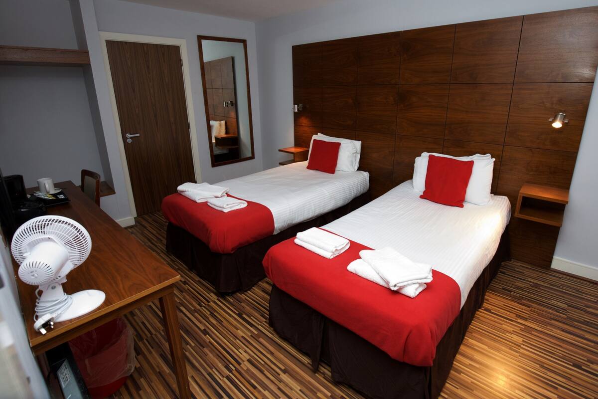 A hotel room with two single beds, a wooden floor, a desk with a fan, and a mirror.