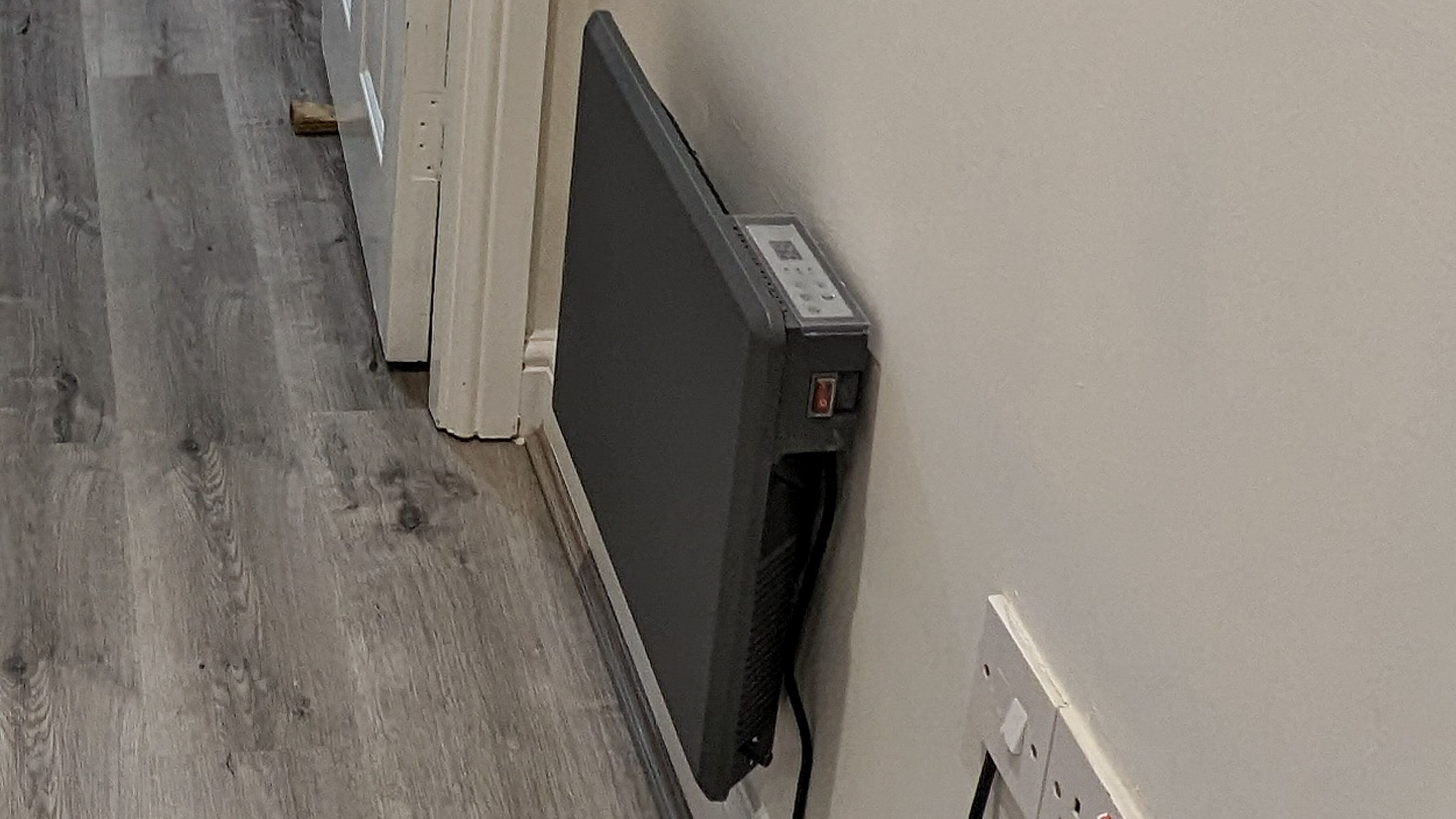 A room with a wooden floor, a white wall, and a black object mounted on the wall.
