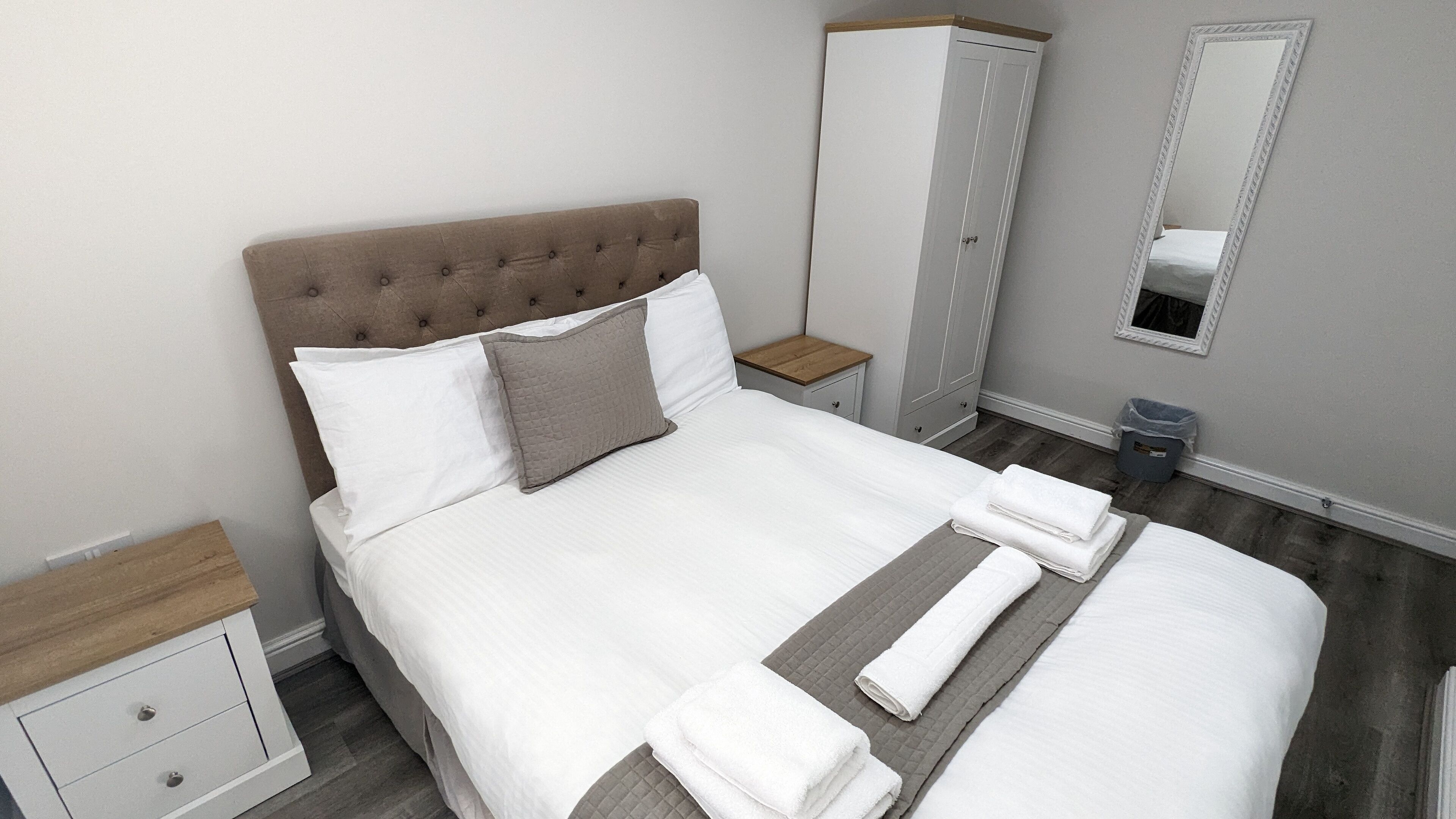 A neatly made bed with white linens and a grey quilted pillow, a wooden nightstand with a drawer, a grey wardrobe, and a mirror on the wall.