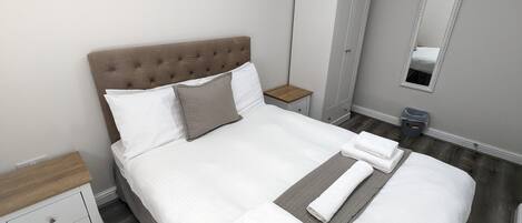 A neatly made bed with white linens and a grey quilted pillow, a wooden nightstand with a drawer, a grey wardrobe, and a mirror on the wall.