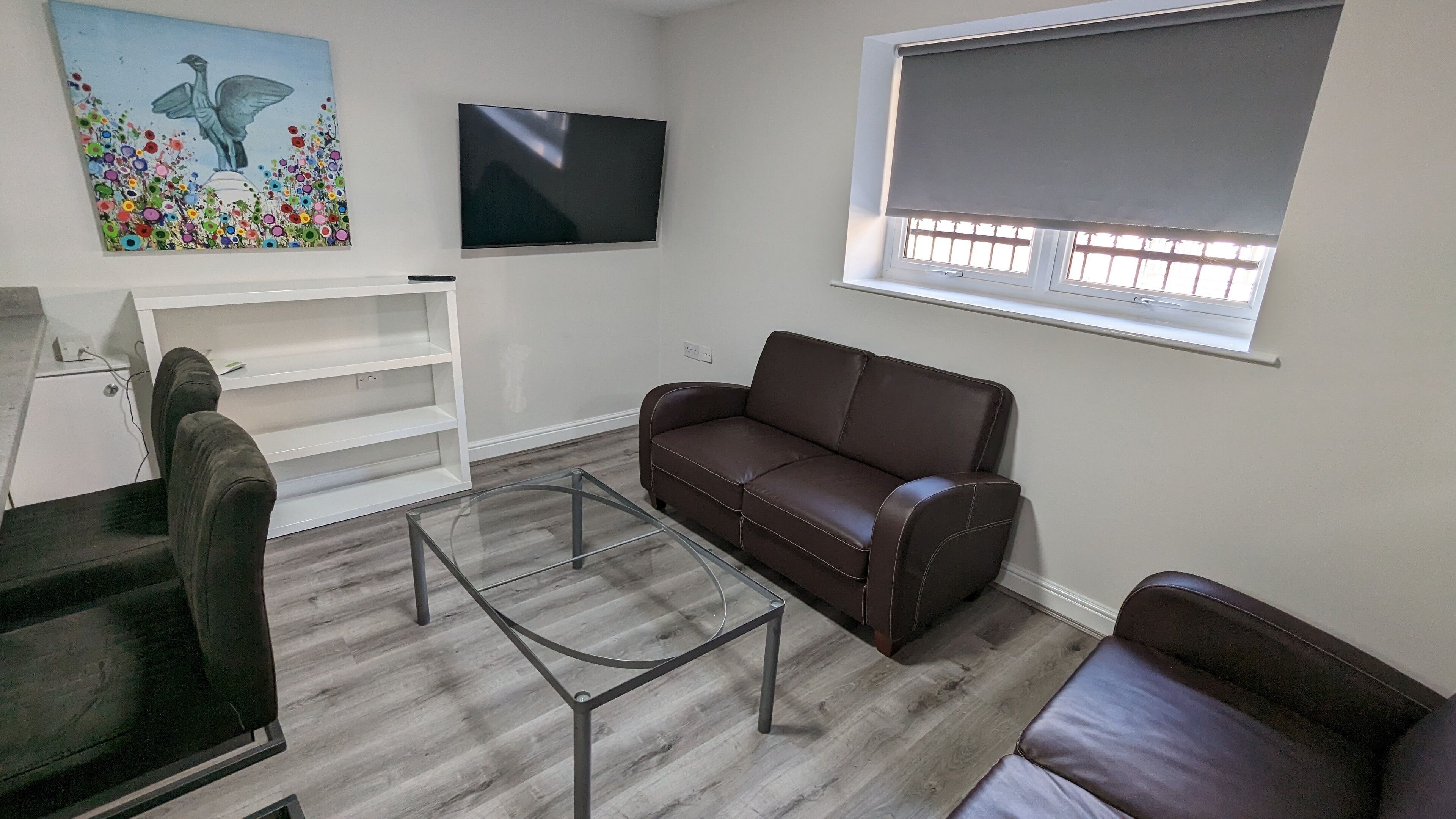 Un salón con un sofá de cuero marrón, una mesa de centro de vidrio, un mueble blanco para la televisión y un cuadro en la pared.
