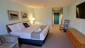 Ein Hotelzimmer mit einem Bett, einem Sessel, einem Schminktisch, einem Fernseher und zwei gerahmten Bildern an der Wand.