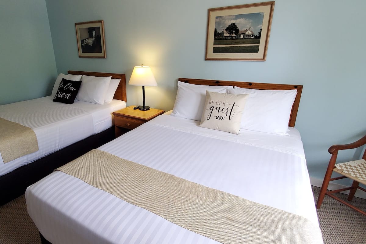 A hotel room with two beds, each with a pillow that says "Be Our Guest," a lamp on one side, and a framed picture on the wall.
