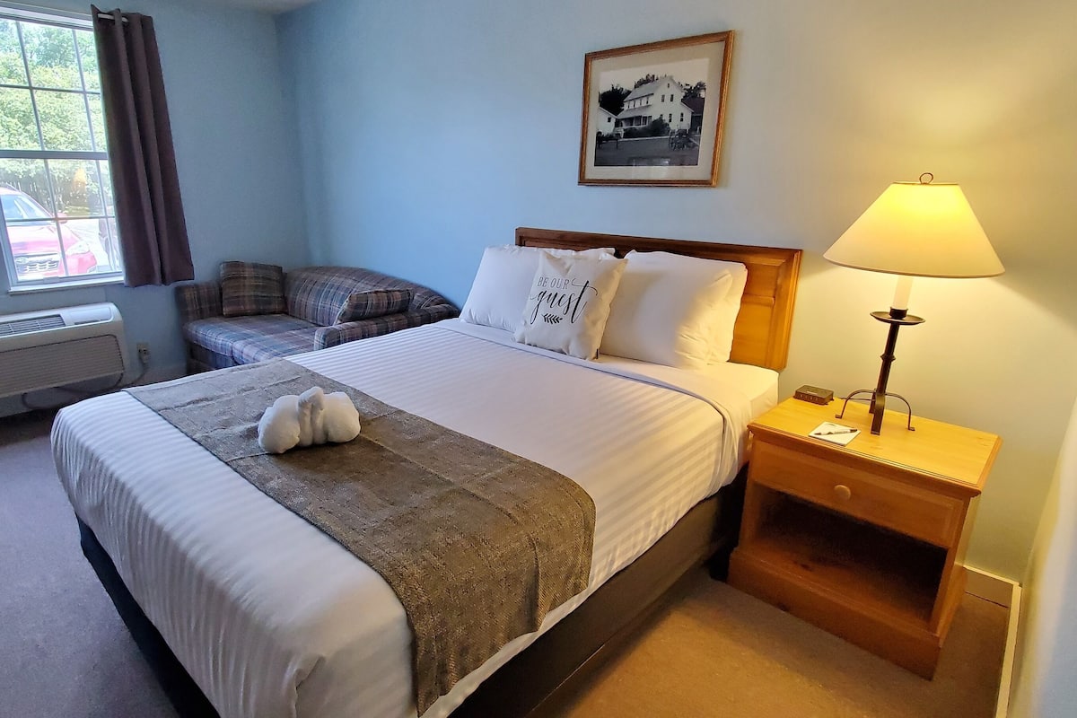 A hotel room with a bed, a bedside table with a lamp, a window with curtains, a wall-mounted picture, and a small heater.