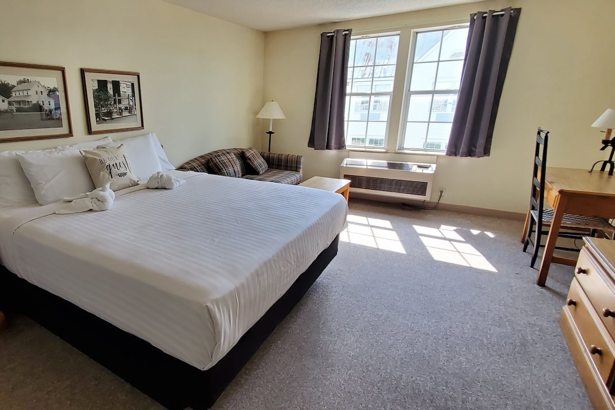 A hotel room with a large bed, a bedside table with a lamp, a desk with a chair, and a window with curtains.