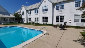 Ein Schwimmbecken mit Metallleiter, eine überdachte Terrasse mit Gartenmöbeln und ein weißes Gebäude mit zahlreichen Fenstern und einem Giebeldach.