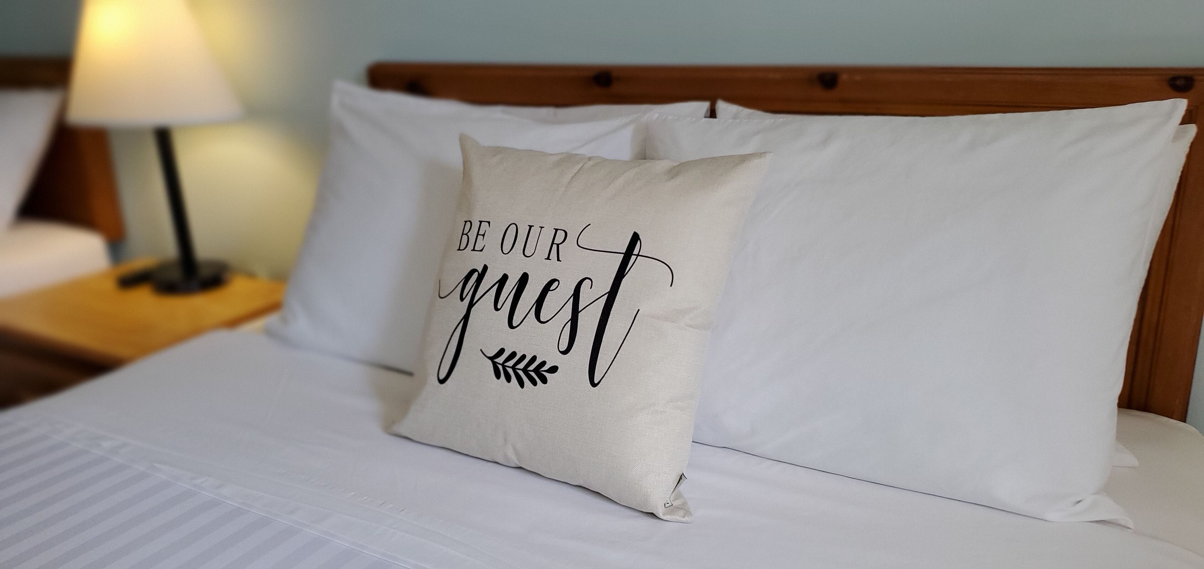 A bed with white bedding and a pillow with the text "BE OUR GUEST" on it.