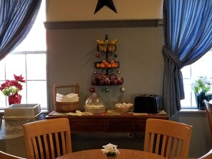 Un comedor con mesa de madera, sillas, una cesta de frutas, un tostador y un frutero en la pared.
