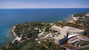 Vue aérienne d’un hôtel situé sur la côte, avec un bâtiment aux lignes courbes, des jardins aménagés et une plage.