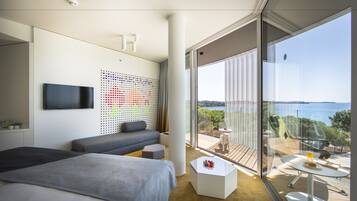 A modern hotel room with a large bed, a sofa, a small table, and a balcony with a view of the sea.