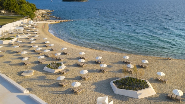 Une plage bordée de rangées de transats et de parasols, un rivage rocheux et une vue sur la mer et les îles lointaines.