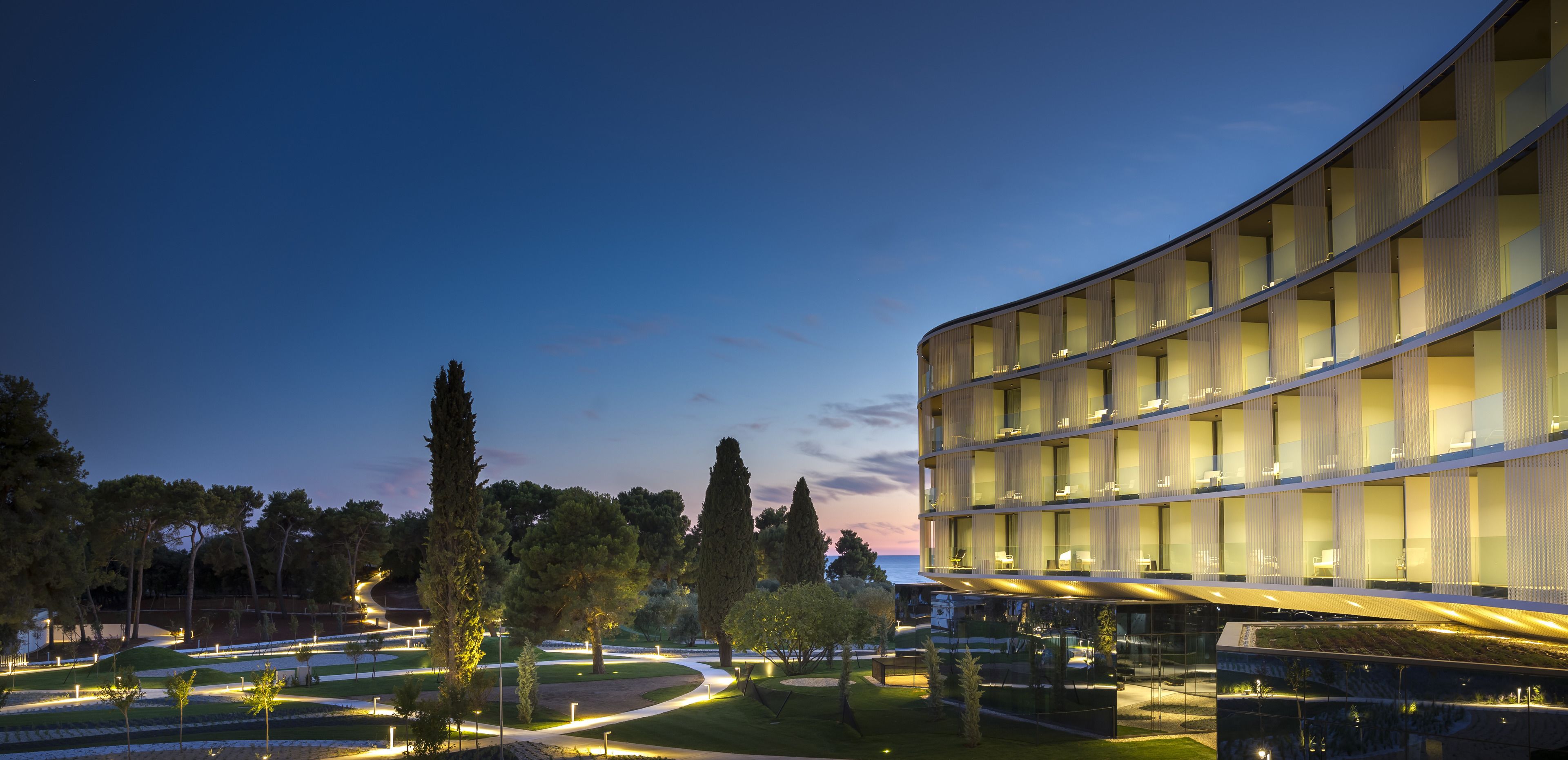 Ein modernes Hotel mit geschwungener Fassade, mehreren Balkonen und einem gut beleuchteten Außenbereich mit Wegen und Bäumen.