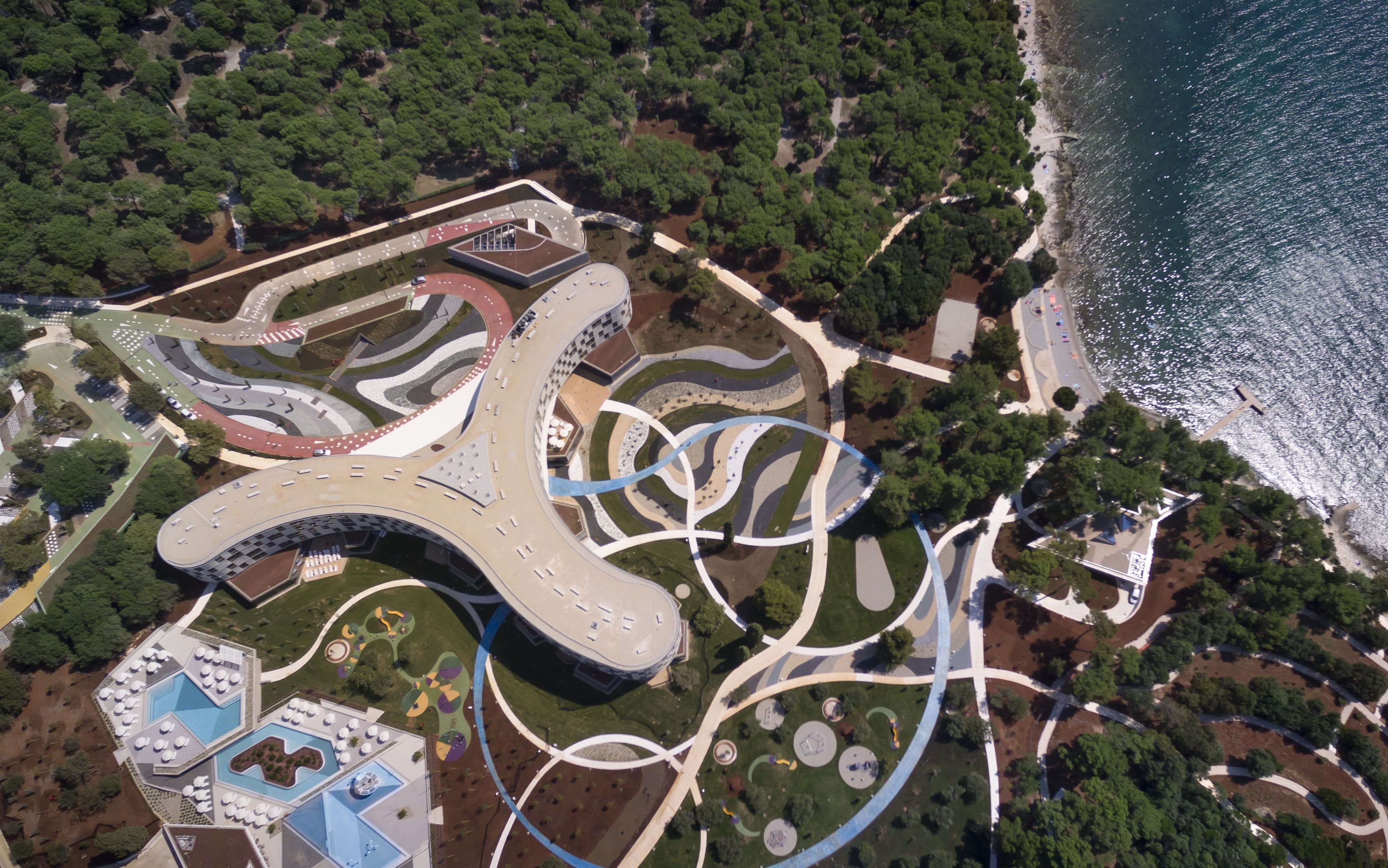 An aerial view of a modern complex with a curved building, landscaped gardens, and a beach.