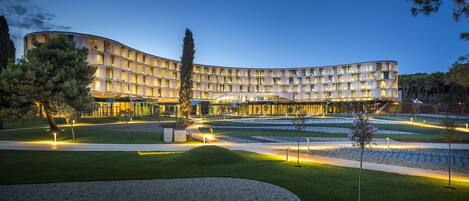 Un hotel moderno con fachada curva, varios balcones y un jardín bien cuidado con senderos y árboles.