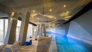 Une piscine intérieure dotée d’un plafond au design unique, de piliers en béton et de chaises longues.